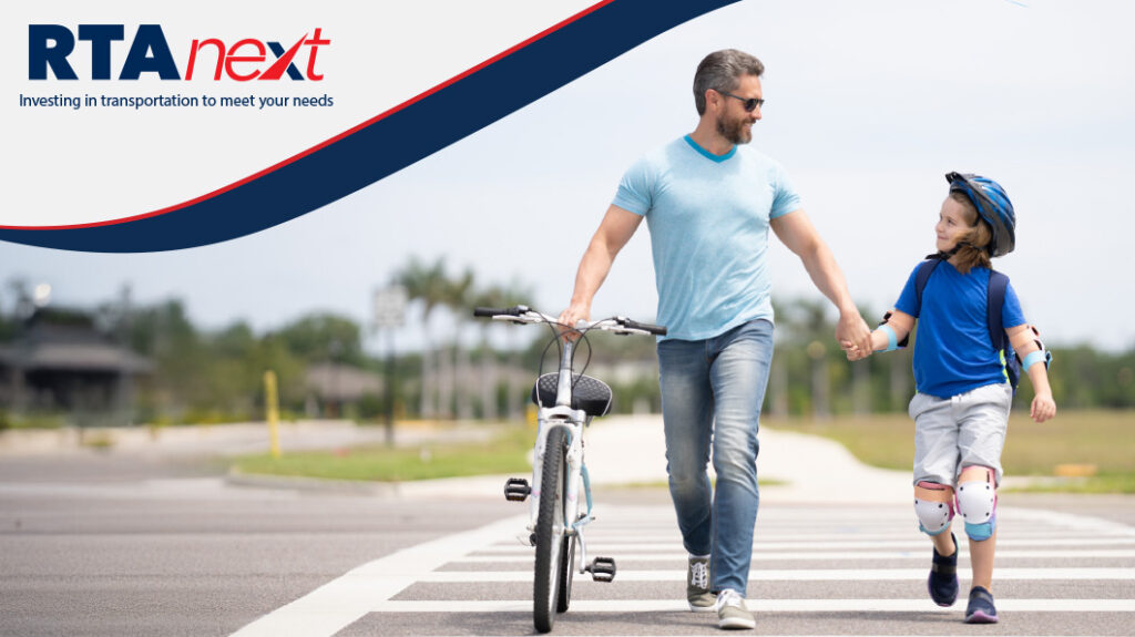 A father leads his daughter hand in hand through a crosswalk while also pushing her bicycle with his other hand.