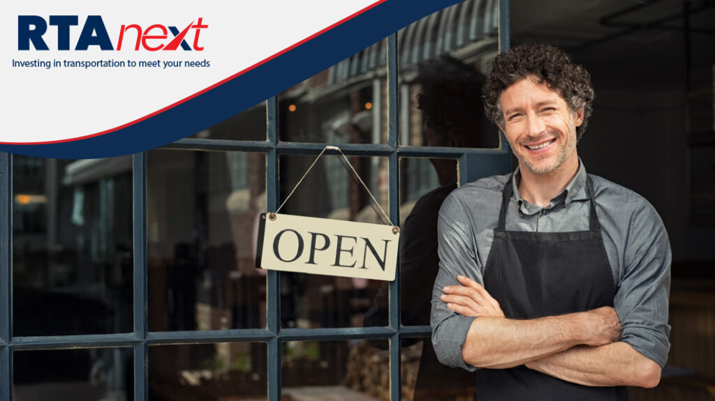 A business owner wearing an apron stands next to an "Open" sign by the entrance.
